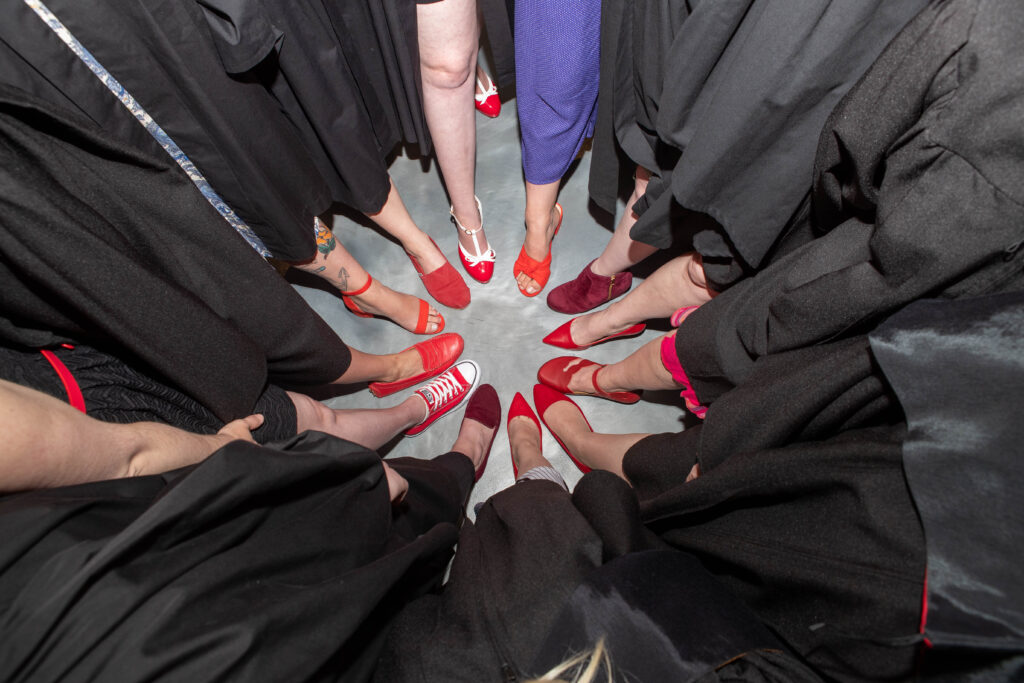 A group of midwifery graduates wearing gowns and red shoes stand with a foot forward making a circle.